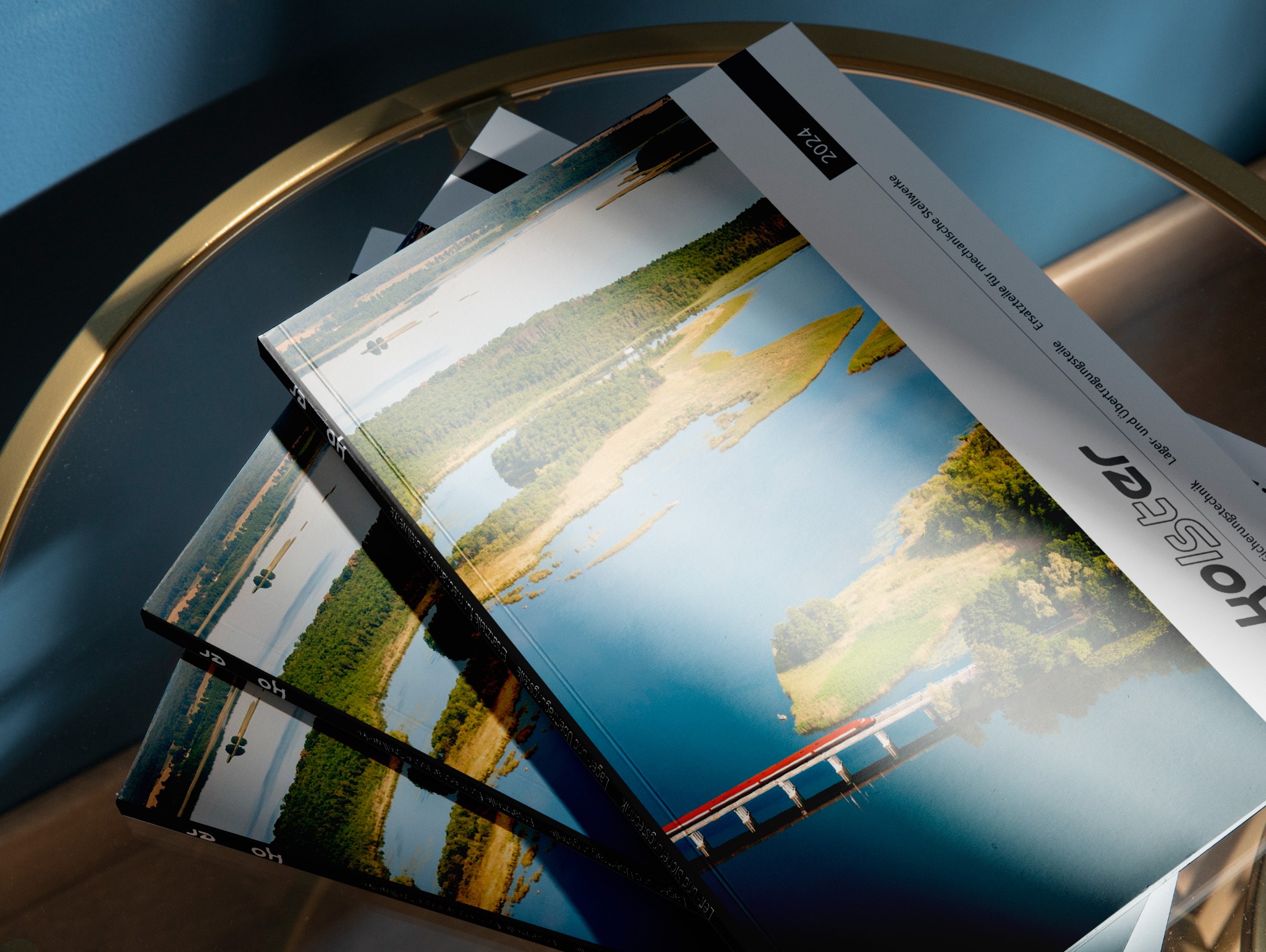 Stos kilku katalogów firmy Holster z widokiem na jezioro i most, leżących na okrągłym, szklanym stoliku z metalową ramą w kolorze złota, na tle niebieskiej ściany.
