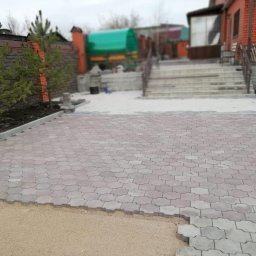 WIKTANA SPÓŁKA Z OGRANICZONĄ ODPOWIEDZIALNOŚCIĄ - Częściowo ułożona kostka brukowa w kształcie fali na podjeździe. Widoczne przygotowane podłoże z piasku i obrzeża. W tle dom z czerwonej cegły i zielone drzewa.