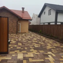 Ułożona kostka brukowa w odcieniach brązu i beżu, tworząca wz&oacute;r na podjeździe. W tle domy: jeden z cegły, drugi otynkowany, oddzielone drewnianym płotem i młodymi drzewkami.