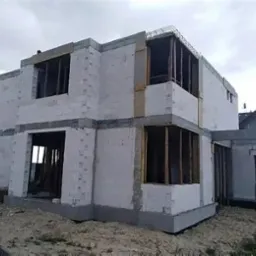 Surowy stan dwupiętrowego domu z betonu komórkowego, widoczne zbrojenia stropu, otwarte okna bez ram, na fundamencie, w tle pochmurne niebo.