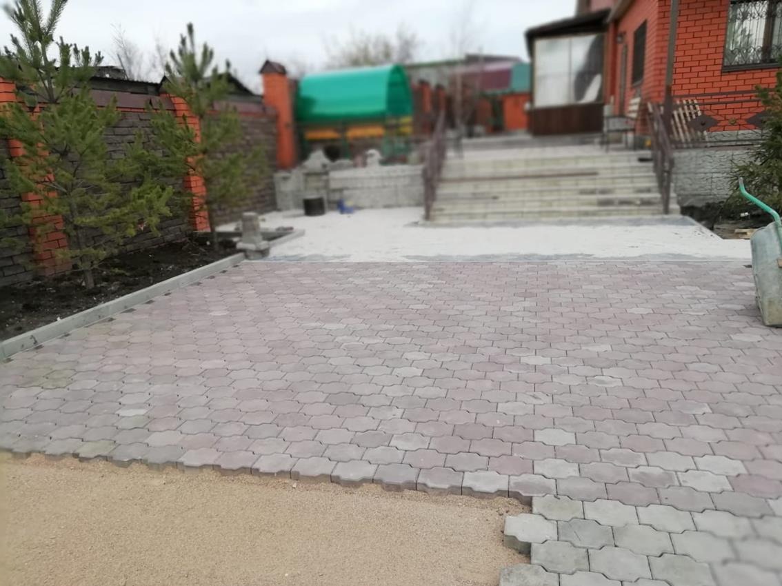 Częściowo ułożona kostka brukowa w kształcie fali na podjeździe. Widoczne przygotowane podłoże z piasku i obrzeża. W tle dom z czerwonej cegły i zielone drzewa.