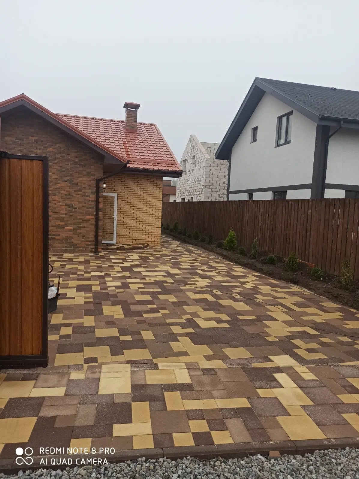 Ułożona kostka brukowa w odcieniach brązu i beżu, tworząca wzór na podjeździe. W tle domy: jeden z cegły, drugi otynkowany, oddzielone drewnianym płotem i młodymi drzewkami.
