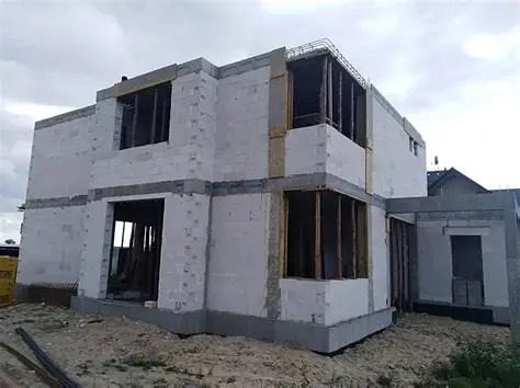 Surowy stan dwupiętrowego domu z betonu komórkowego, widoczne zbrojenia stropu, otwarte okna bez ram, na fundamencie, w tle pochmurne niebo.