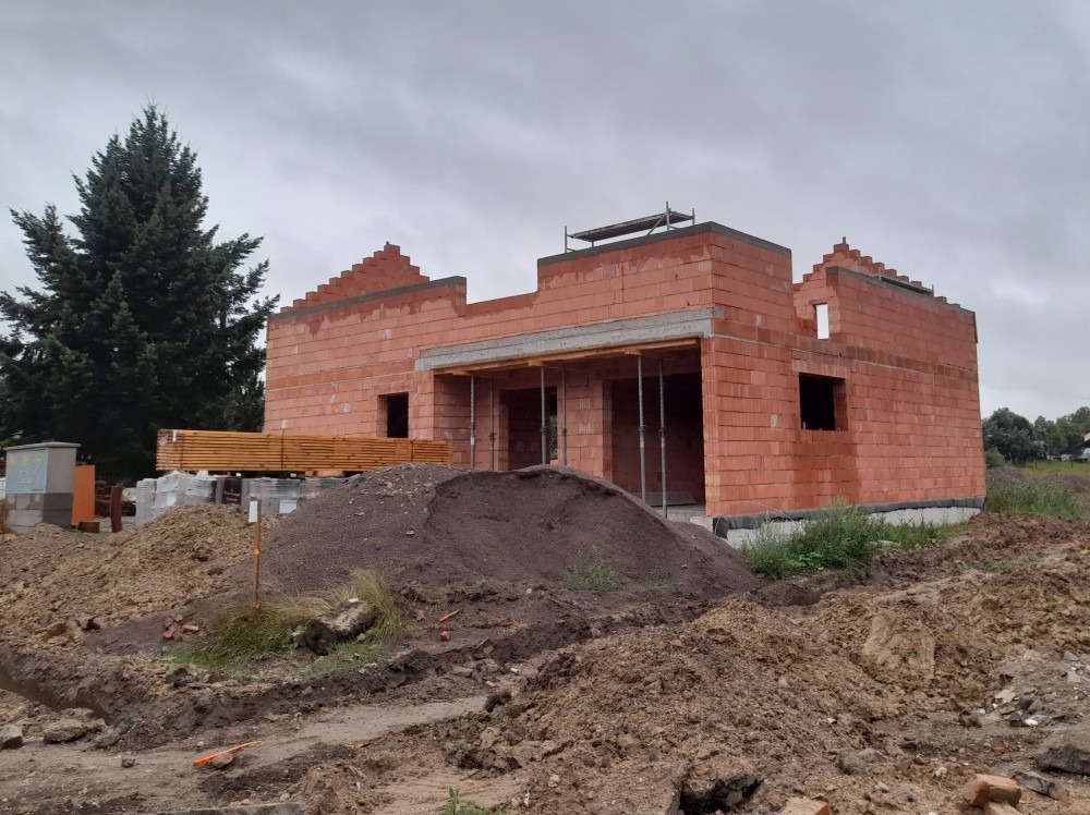 Surowy stan domu z czerwonej cegły, widoczne drewniane elementy konstrukcyjne i rusztowanie, w tle iglaste drzewo i pochmurne niebo. Teren budowy z hałdami ziemi.