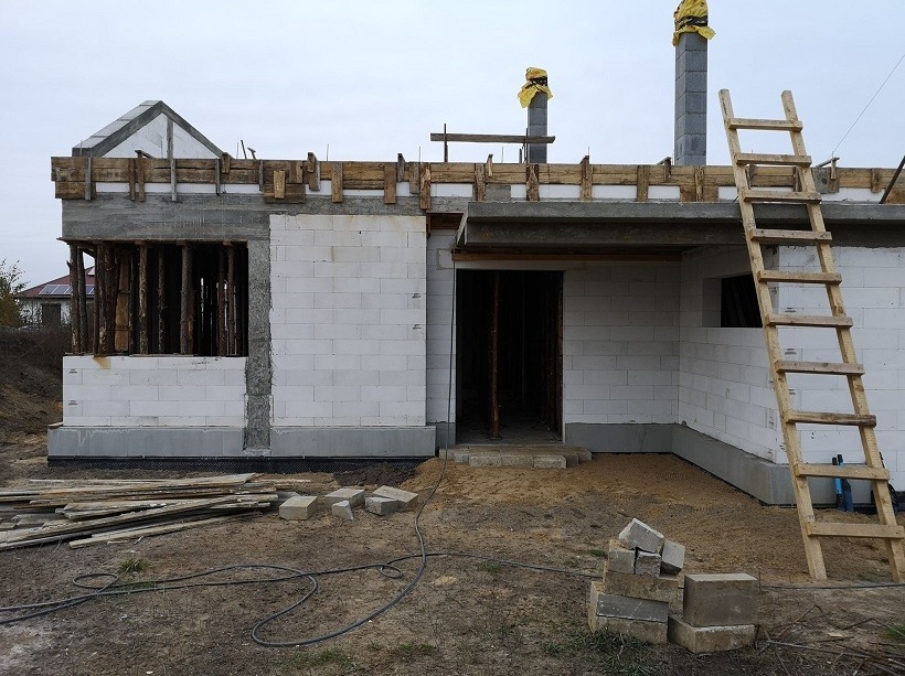 Wznoszenie ścian domu z białych bloczków, widoczne drewniane szalunki i podpory. Na pierwszym planie ziemia, cegły i kabel elektryczny. Drabina oparta o ścianę.