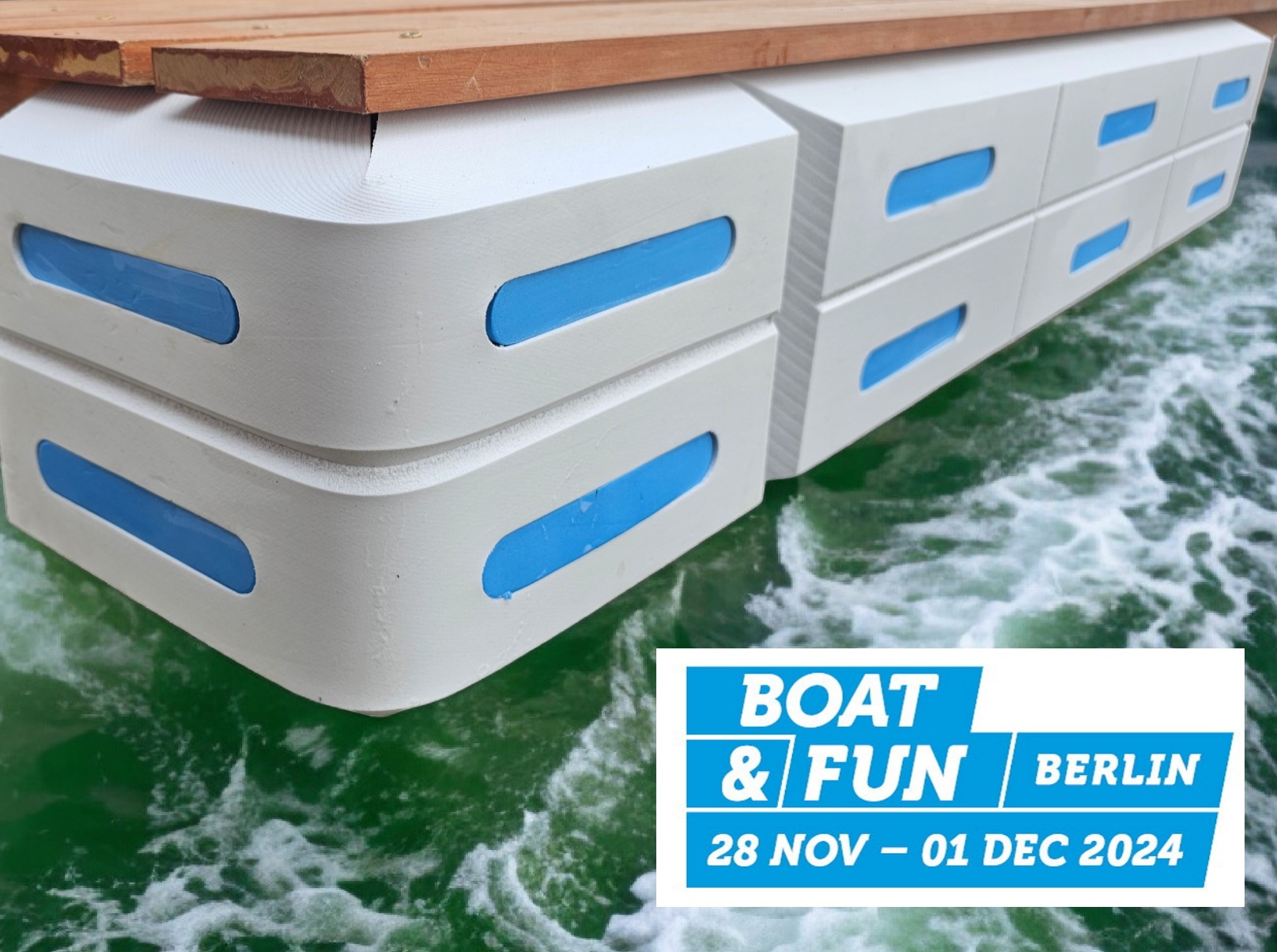Białe, modułowe pływaki z niebieskimi akcentami, pod drewnianym pomostem, unoszące się na wzburzonej wodzie. Widoczne logo targów Boat & Fun Berlin.
