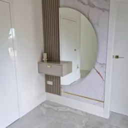 Nowoczesna aranżacja przedpokoju z okrągłym lustrem w marmurowej ramie, drewnianymi lamelami i szarą szafką z ozdobnym uchwytem. Minimalistyczny design.