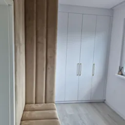 Nowoczesna zabudowa wnęki z tapicerowanym siedziskiem i panelem ściennym w odcieniach beżu, dopasowana do białej szafy z podłużnymi uchwytami i jasnej podłogi.