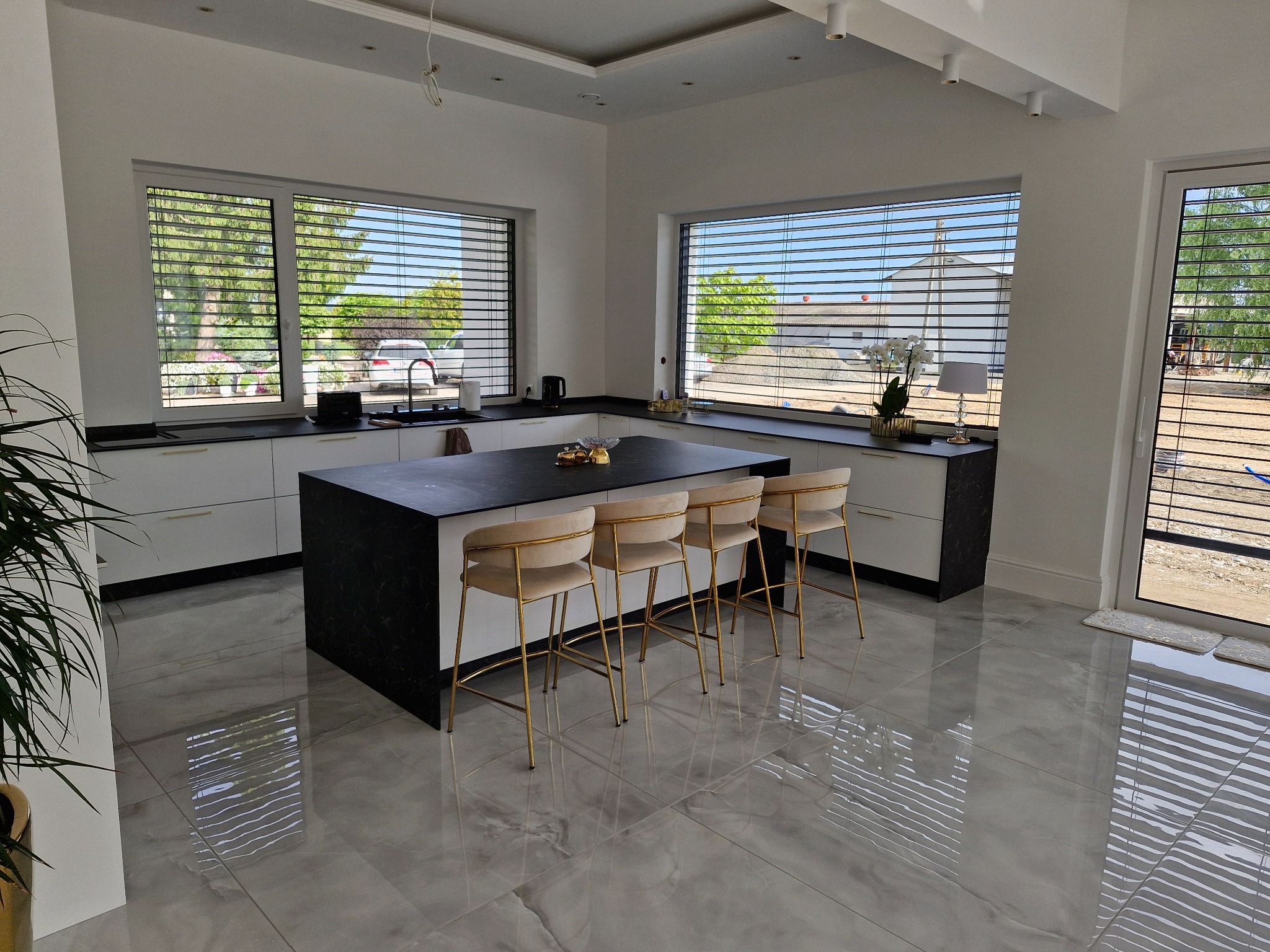 Nowoczesna kuchnia z wyspą i białymi szafkami, czarny blat, złote krzesła barowe, duże okna z żaluzjami, marmurowe płytki podłogowe.