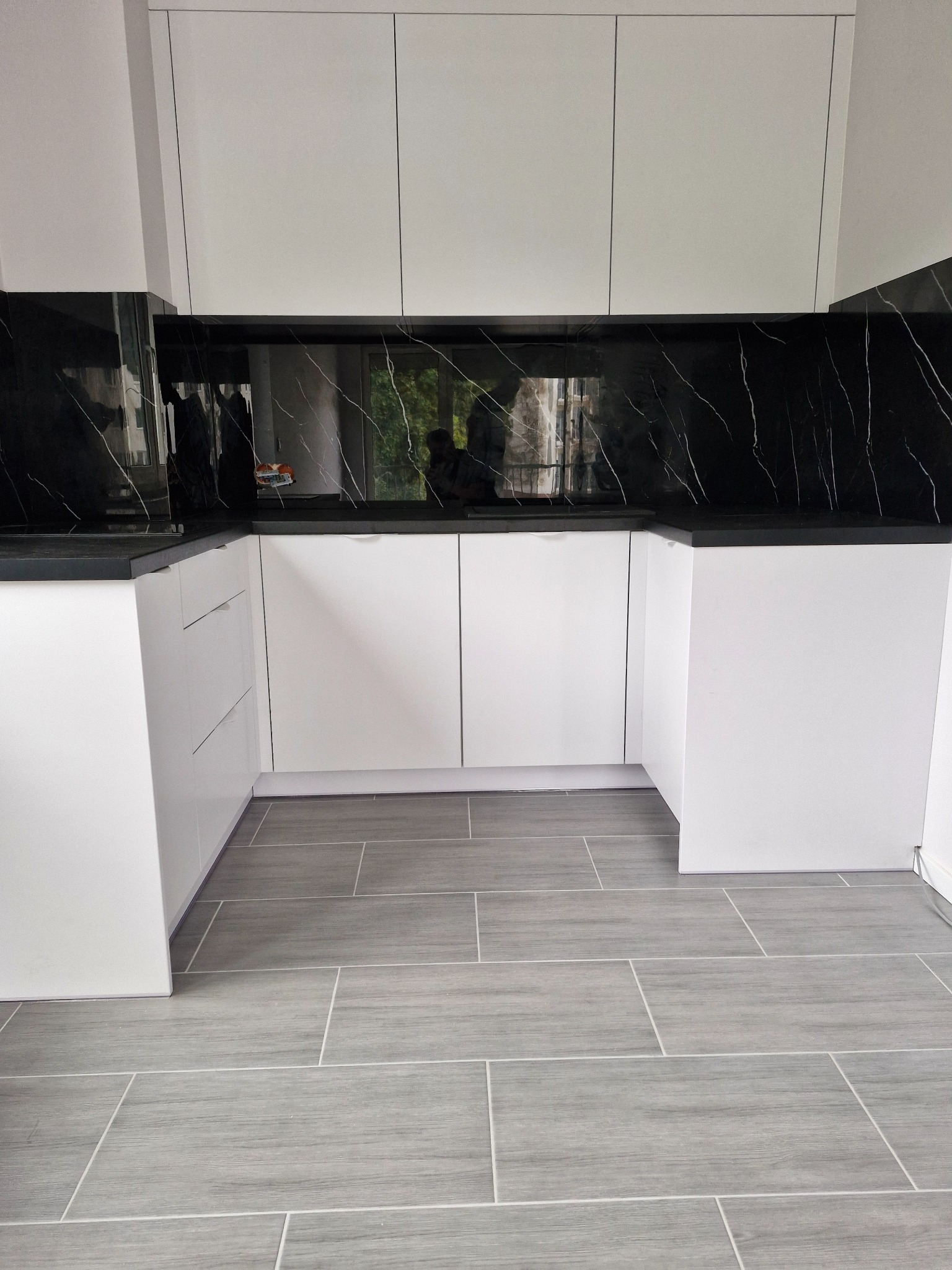 Nowoczesna kuchnia na wymiar z białymi frontami i czarnym blatem w Korfantowie. Minimalistyczny design, szare płytki podłogowe, odbicie w panelu nad blatem.