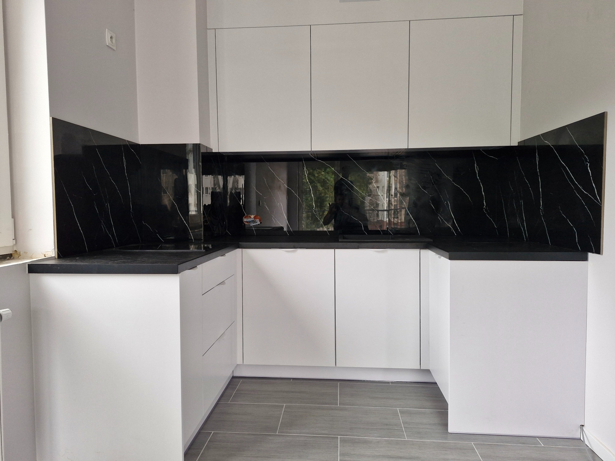 Nowoczesna, biała kuchnia z czarnym blatem i panelem imitującym marmur. Minimalistyczny design, szare płytki podłogowe, widok z przodu.