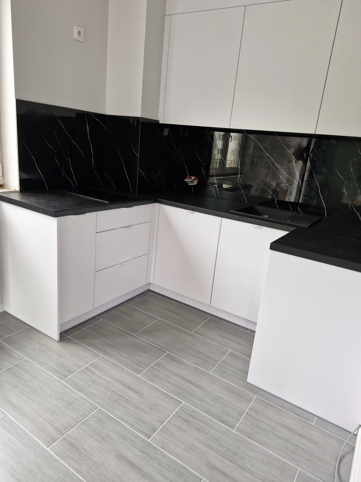 Nowoczesna kuchnia z białymi szafkami i czarnym blatem imitującym marmur. Szare płytki podłogowe. Minimalistyczny design, widok z niskiego kąta.