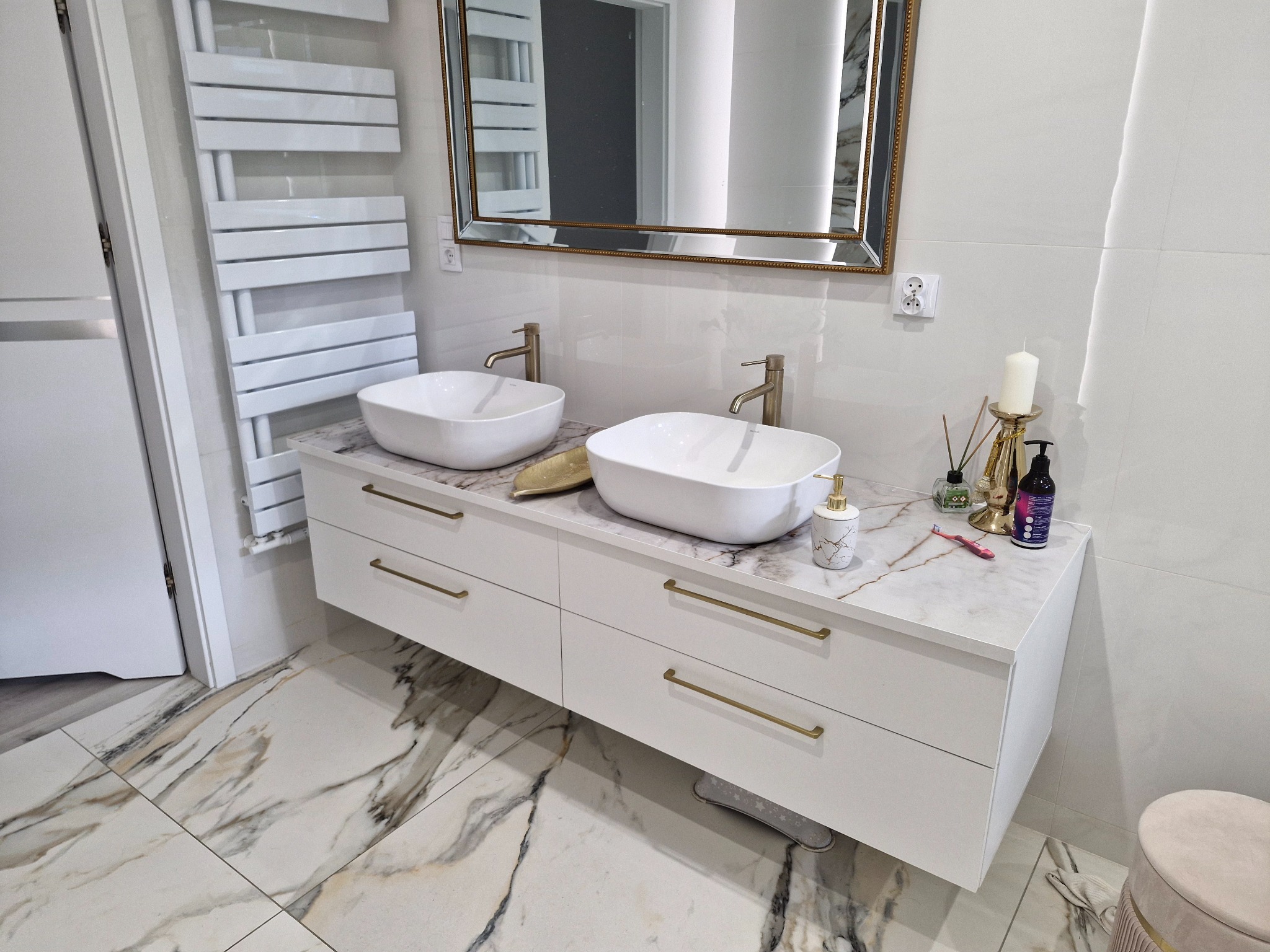 Biała szafka łazienkowa z marmurowym blatem i dwiema umywalkami nablatowymi, złote detale, płytki podłogowe w marmurowy wzór, nowoczesny design.