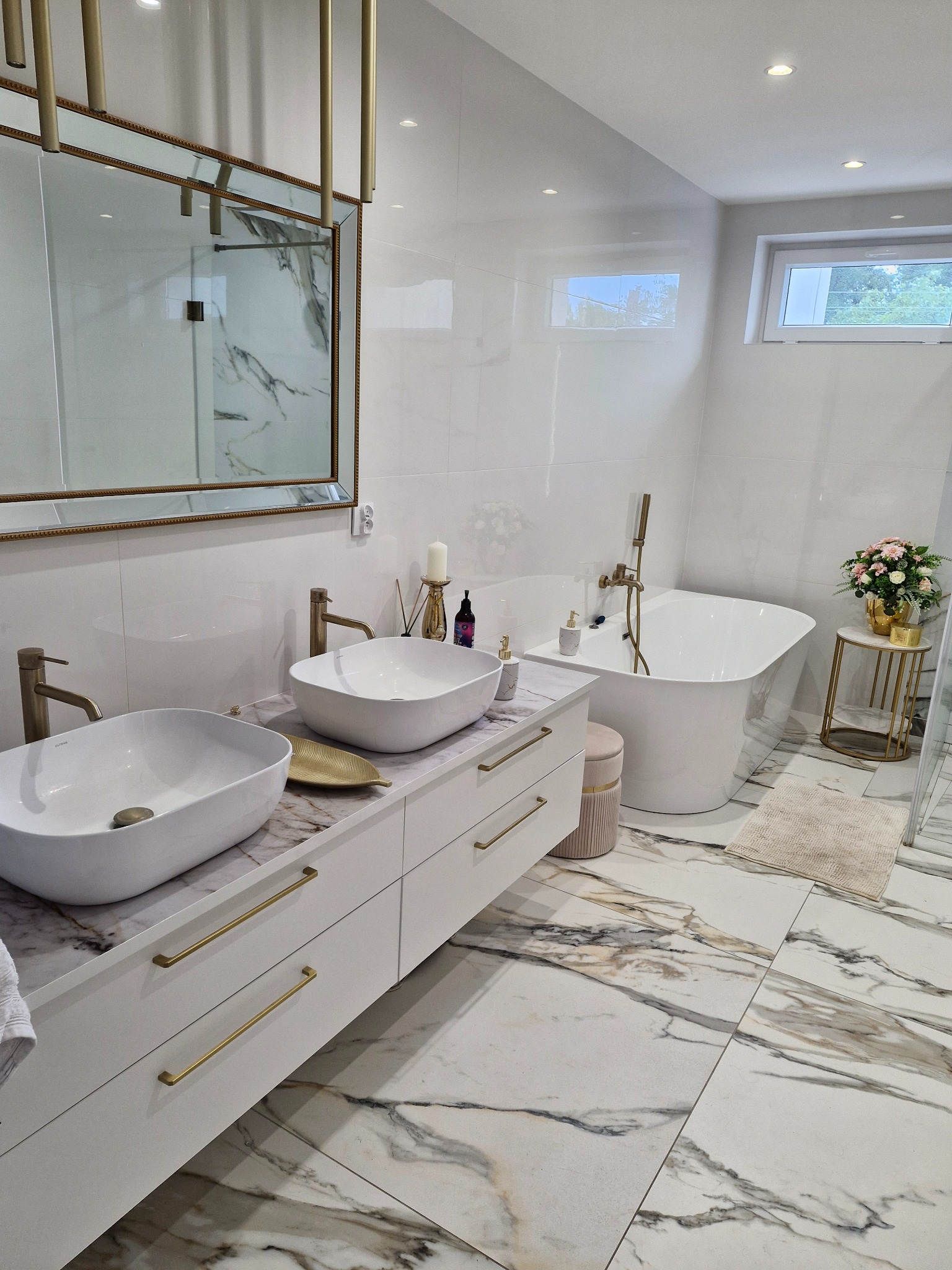 Elegancka łazienka z marmurową podłogą, białymi szafkami z złotymi uchwytami, owalnymi umywalkami i wolnostojącą wanną. Lustro z dekoracyjną ramą.