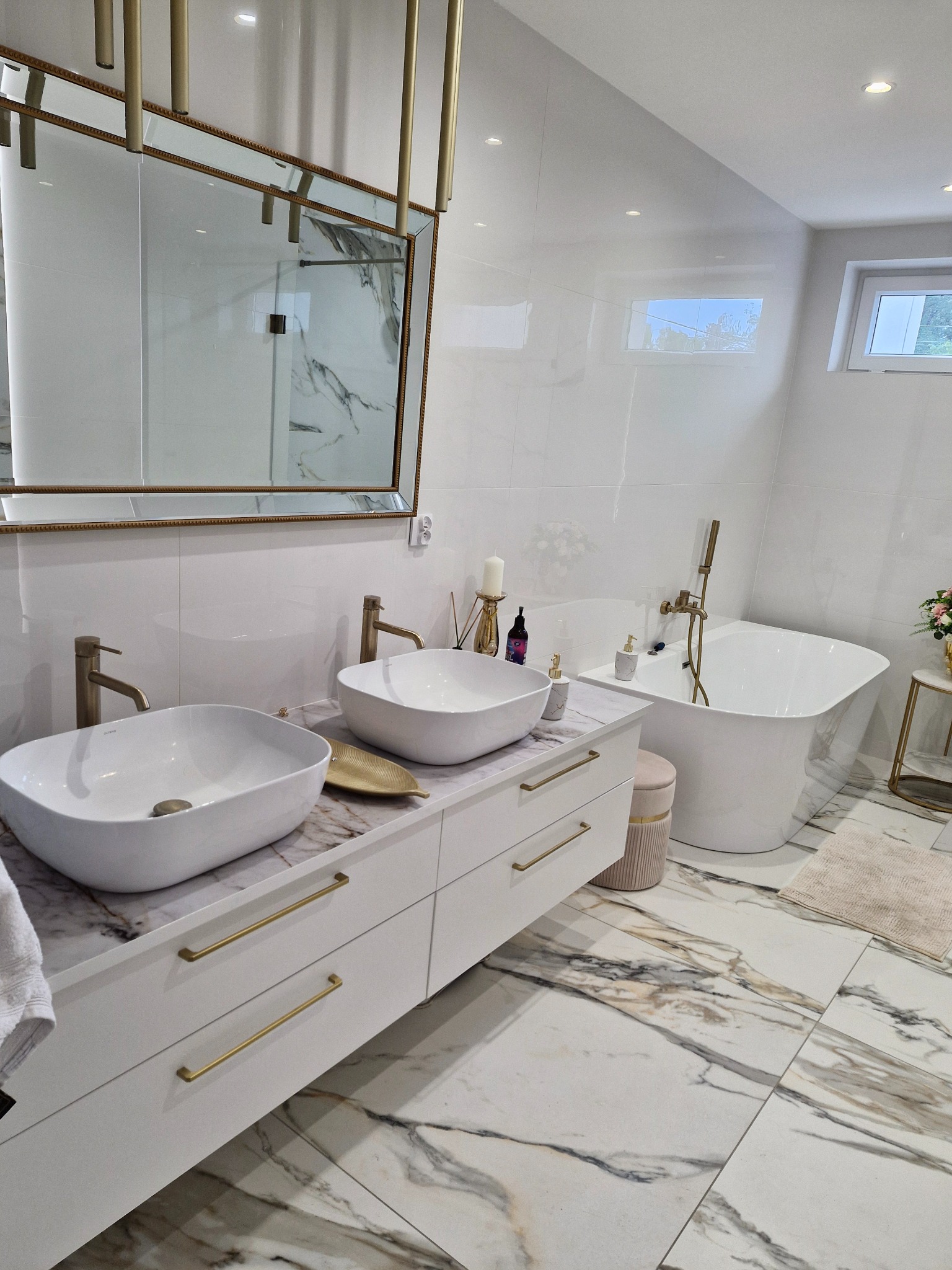 Jasna łazienka z marmurową podłogą, białymi meblami z złotymi uchwytami, dwiema umywalkami i wanną. Złote akcenty w armaturze i lustrze.