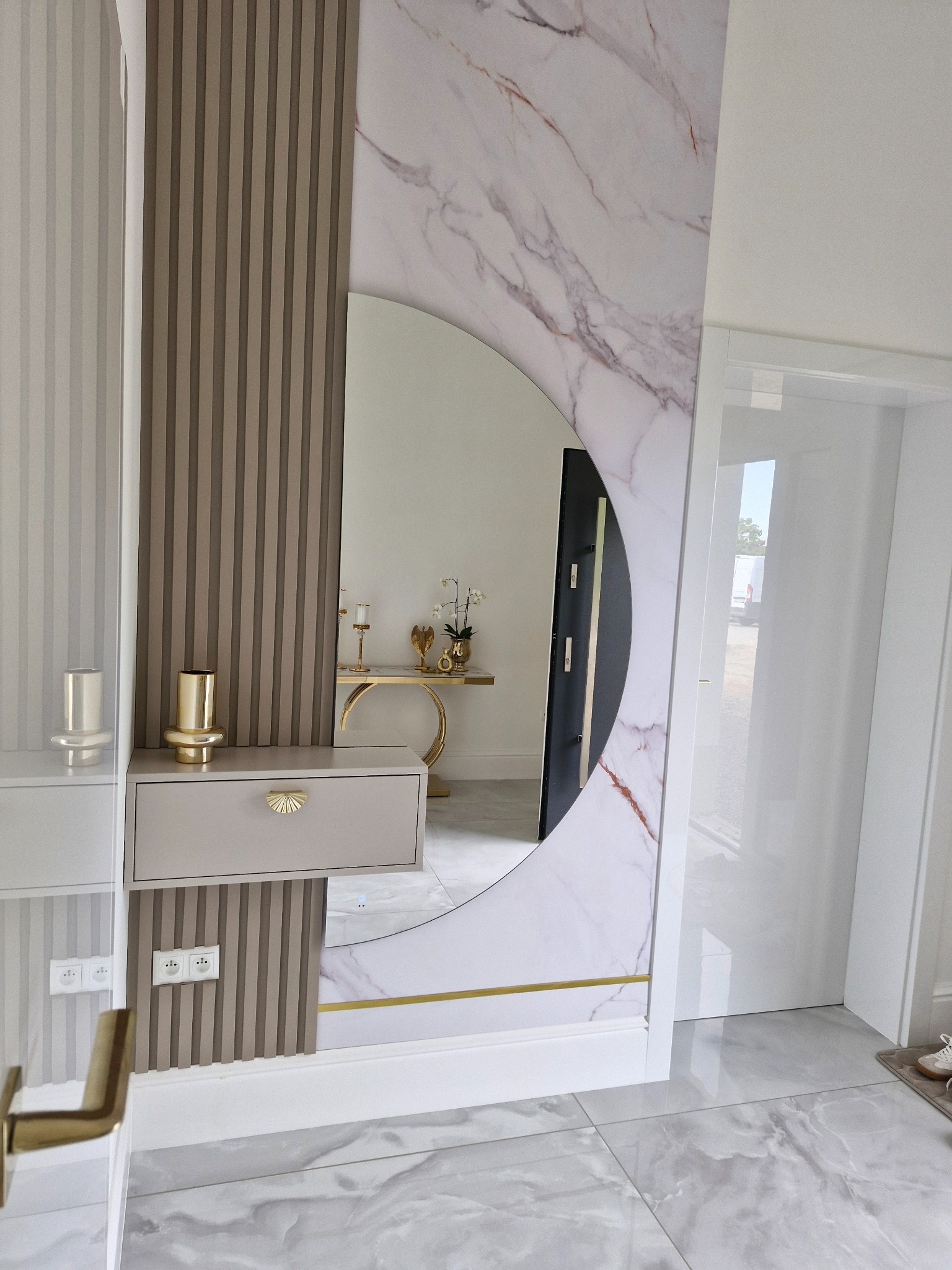 Nowoczesny przedpokój z marmurową ścianą, półokrągłym lustrem i beżową szafką z dekoracyjnym uchwytem. Widoczne gniazdka elektryczne i złote akcenty dekoracyjne.