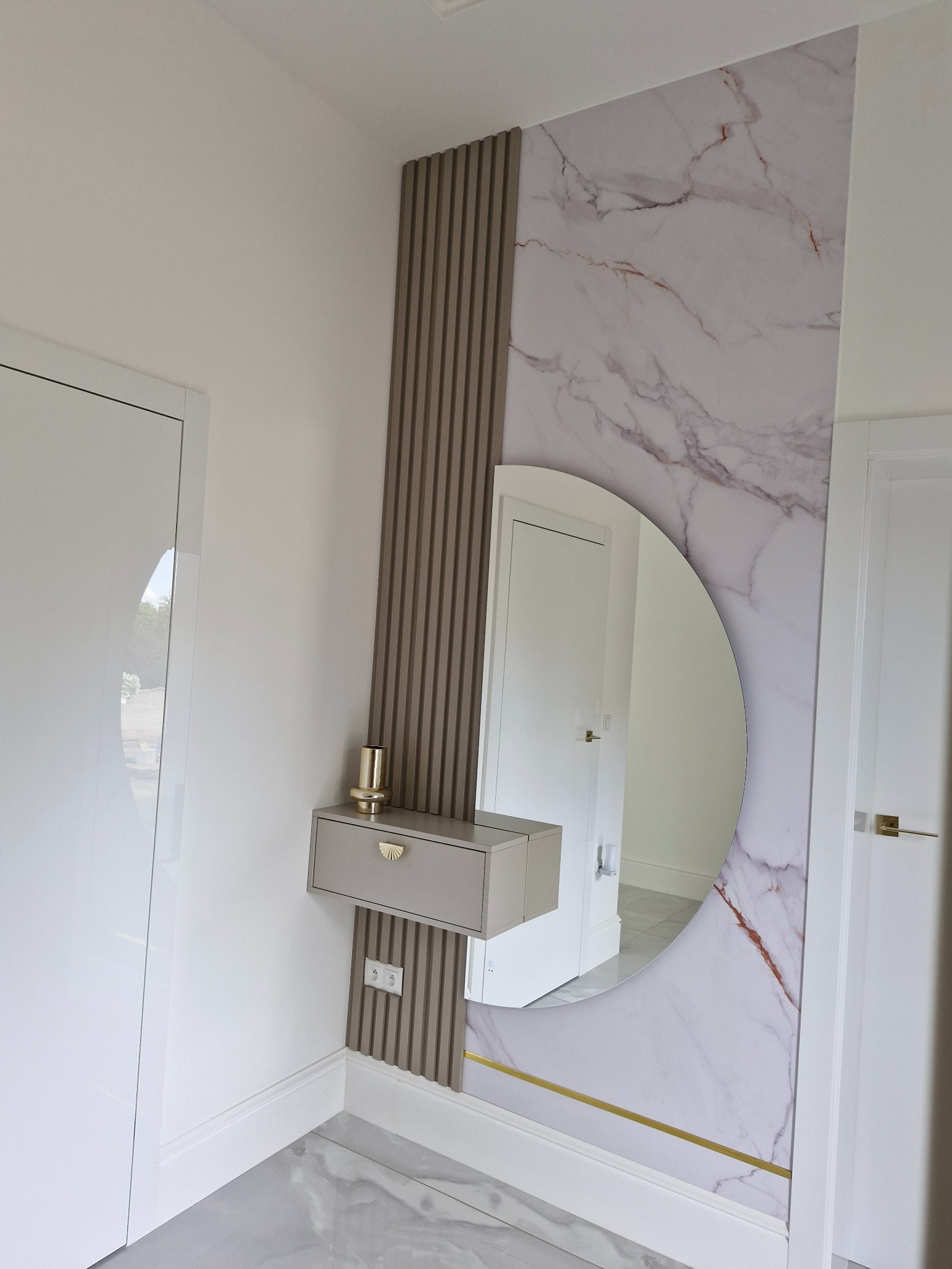 Nowoczesna aranżacja przedpokoju: okrągłe lustro, marmurowa tapeta, pionowe lamele i wisząca szafka z dekoracyjnym uchwytem. Widoczne gniazdka elektryczne i połyskująca podłoga.