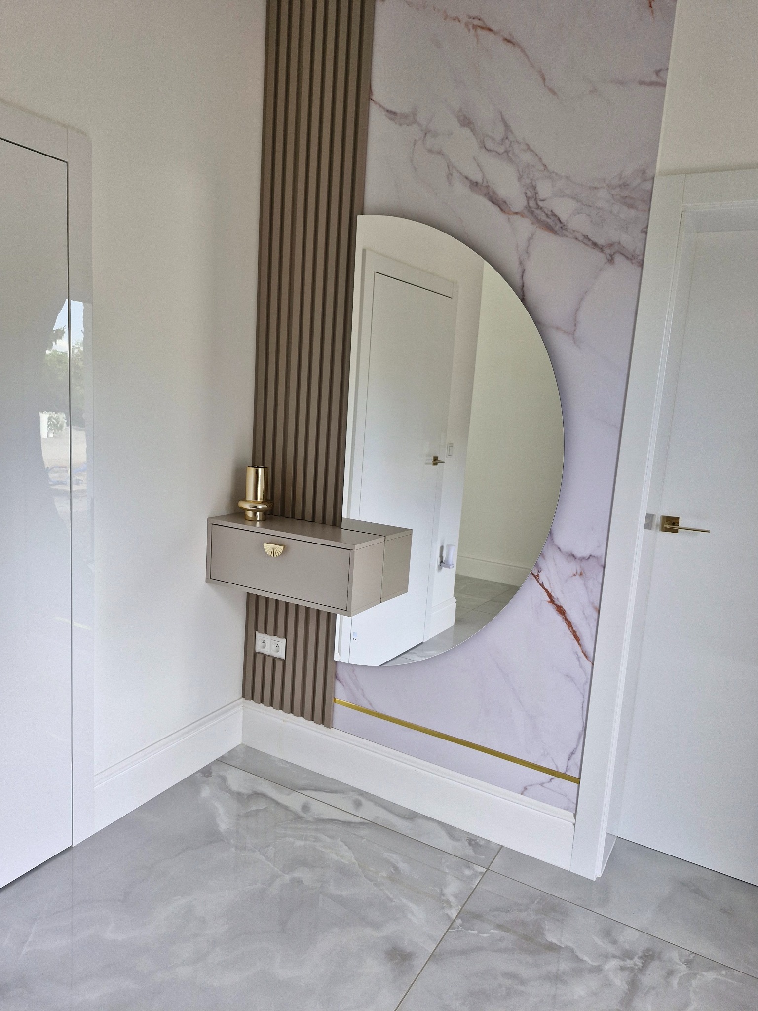 Nowoczesna aranżacja przedpokoju: okrągłe lustro z marmurowym tłem, pionowe lamele, pływająca szuflada z złotym akcentem i kontakt elektryczny. Minimalistyczny design.