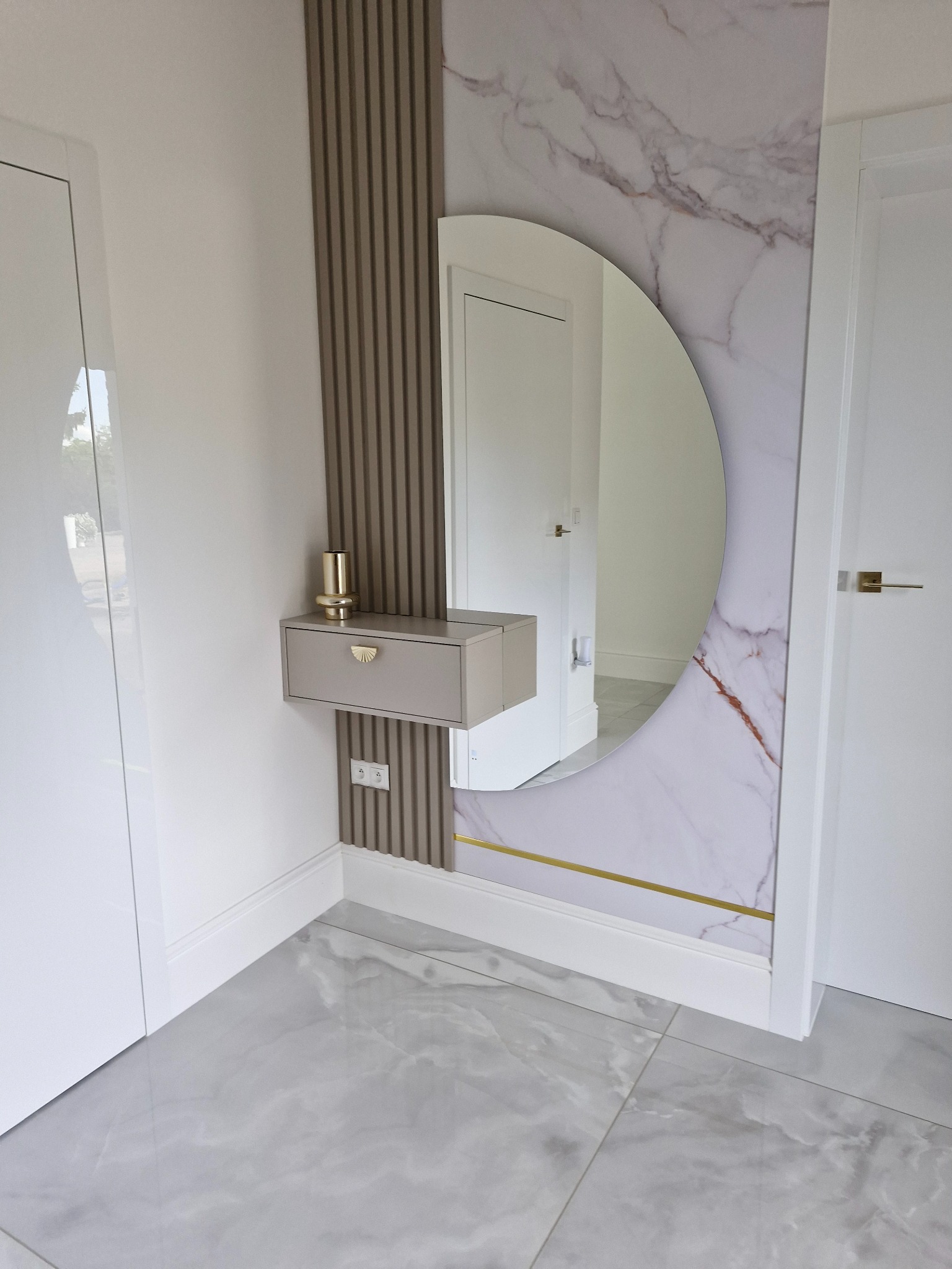 Nowoczesna aranżacja przedpokoju z okrągłym lustrem w marmurowej ramie, drewnianymi lamelami i szarą szafką z ozdobnym uchwytem. Minimalistyczny design.