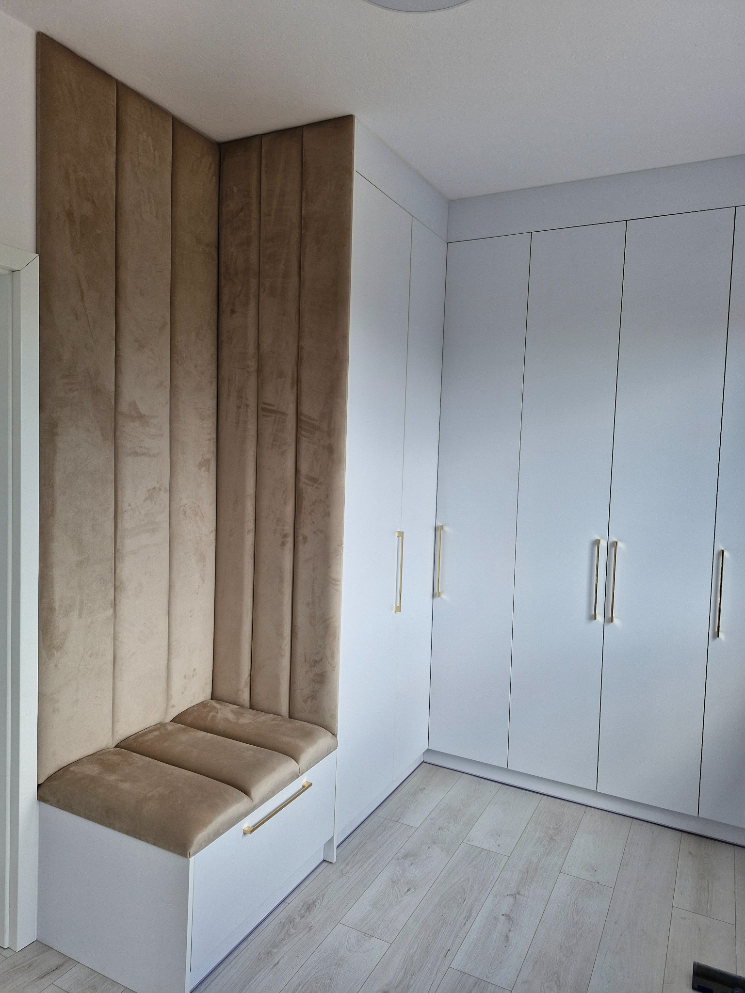 Biała szafa wnękowa z siedziskiem obitym beżową tkaniną i złotymi uchwytami, tworząca spójną zabudowę w jasnym wnętrzu.