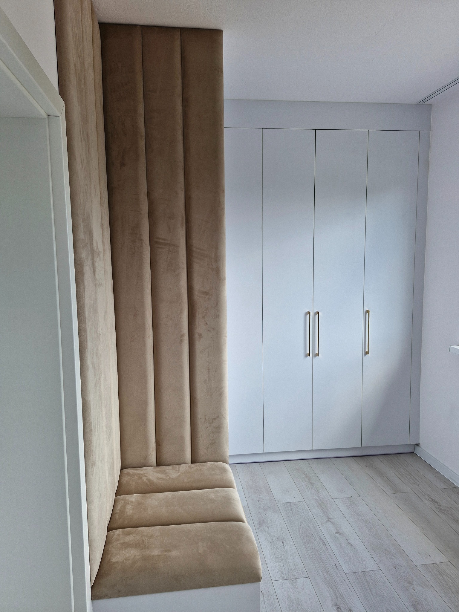 Beżowa, tapicerowana ściana z siedziskiem w nowoczesnym wnętrzu. W tle biała szafa wnękowa z złotymi uchwytami i jasna podłoga. Ujęcie z perspektywy podłogi.