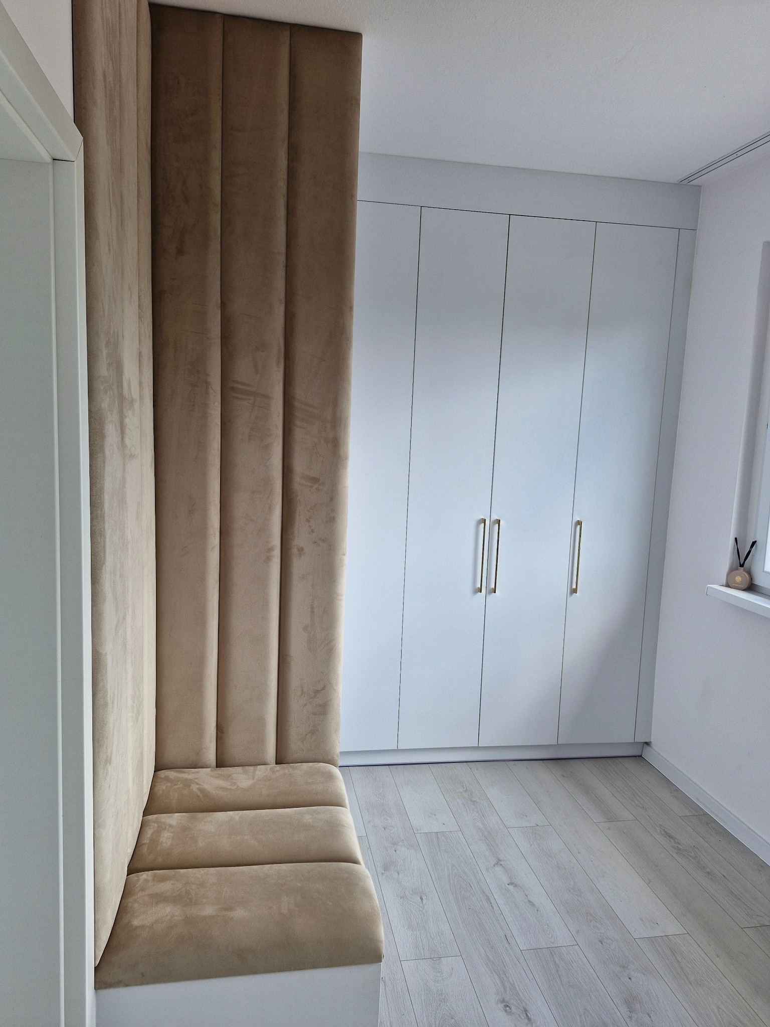 Nowoczesna zabudowa wnęki z tapicerowanym siedziskiem i panelem ściennym w odcieniach beżu, dopasowana do białej szafy z podłużnymi uchwytami i jasnej podłogi.
