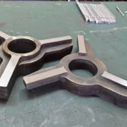 Dwa metalowe, symetryczne elementy z otworem centralnym i trzema ramionami, leżące na zielonym stole warsztatowym obok narzędzi. Obróbka metalu.