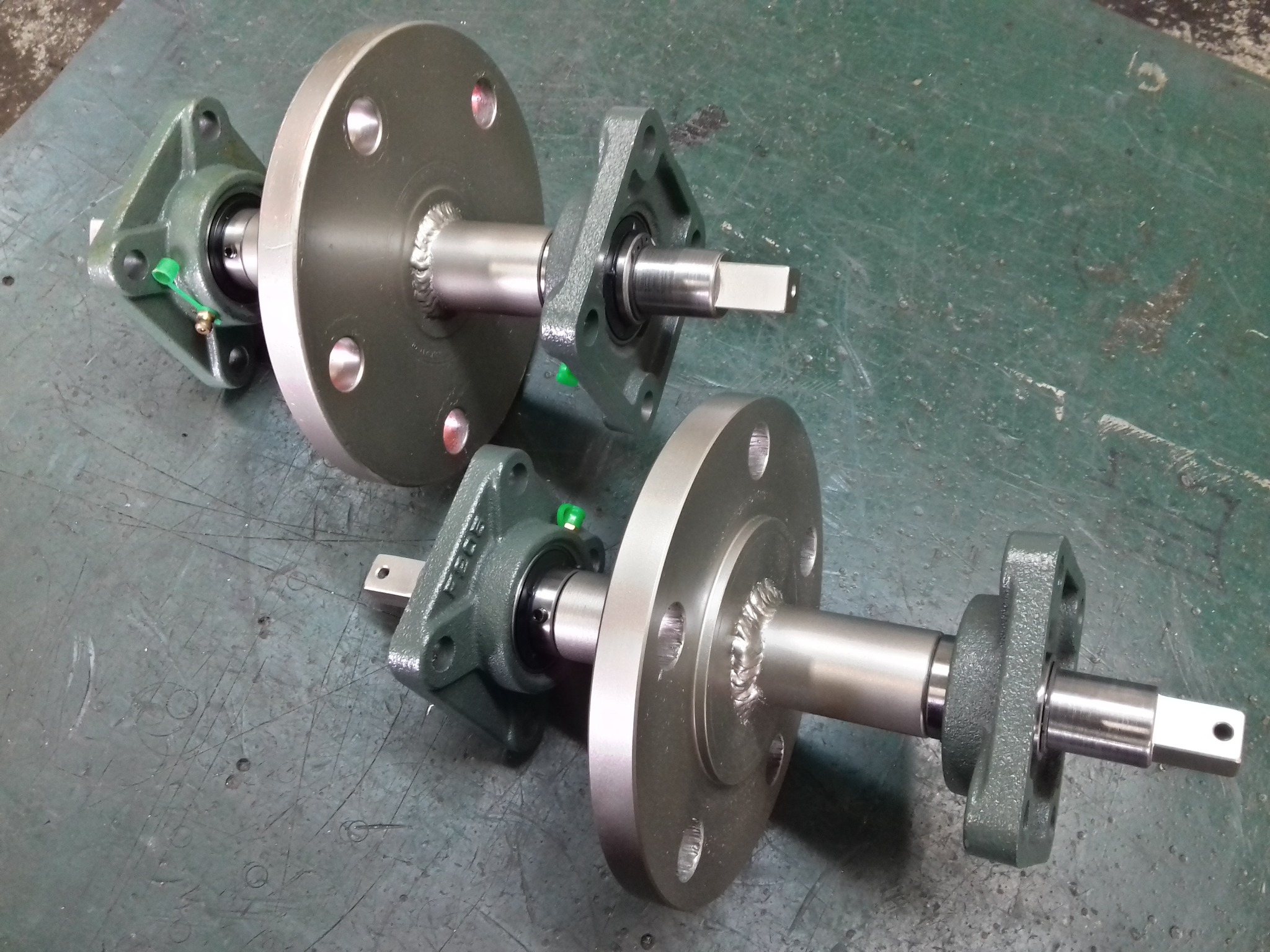 Dwa wały z łożyskami i kołnierzami, połączone spawem, leżą na zielonej, zarysowanej powierzchni. Widoczne detale obróbki i mocowania.