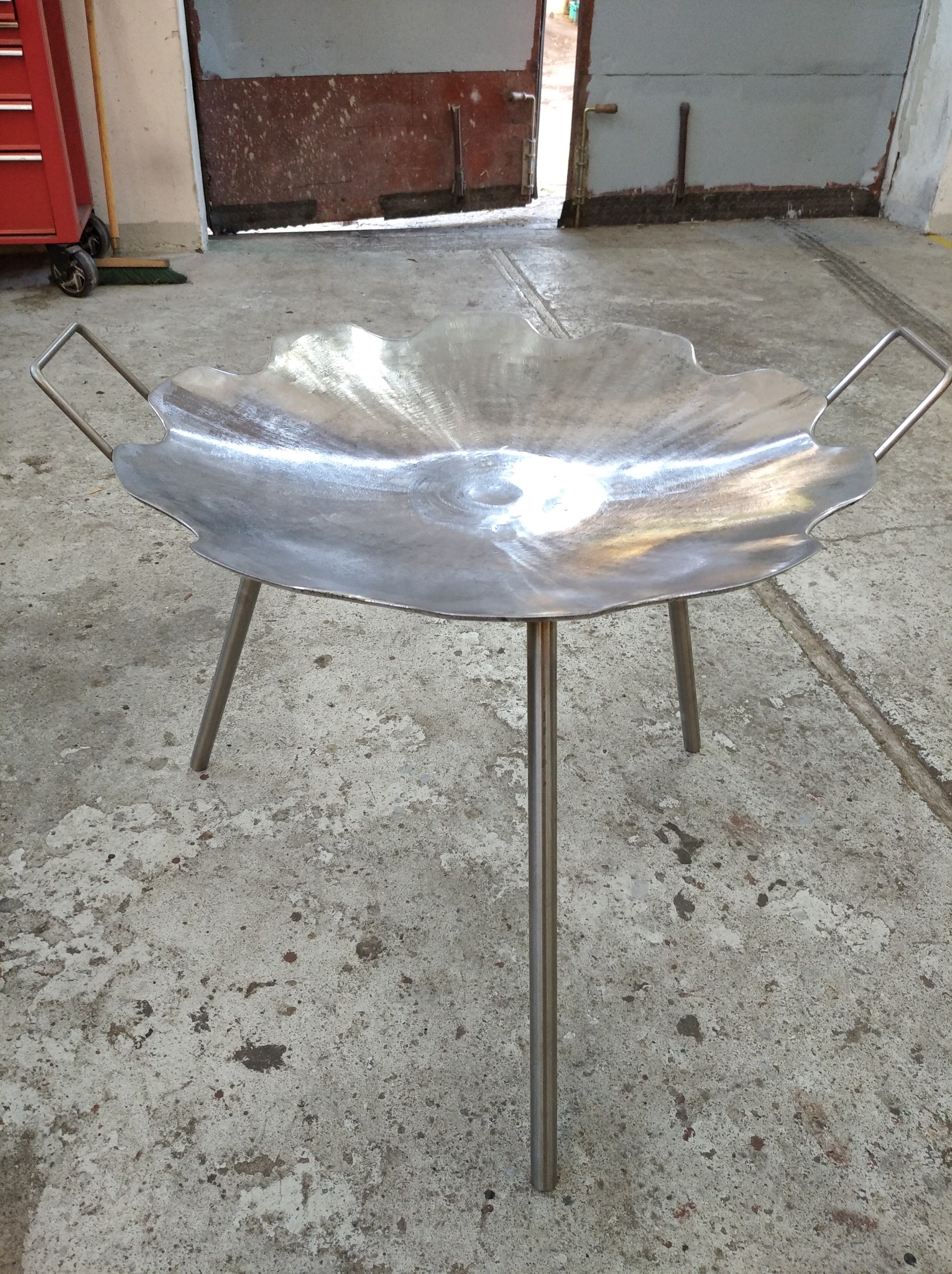Metalowa misa na trójnogu z uchwytami, przypominająca palenisko, stoi na betonowej posadzce w warsztacie. Widoczne ślady obróbki metalu.