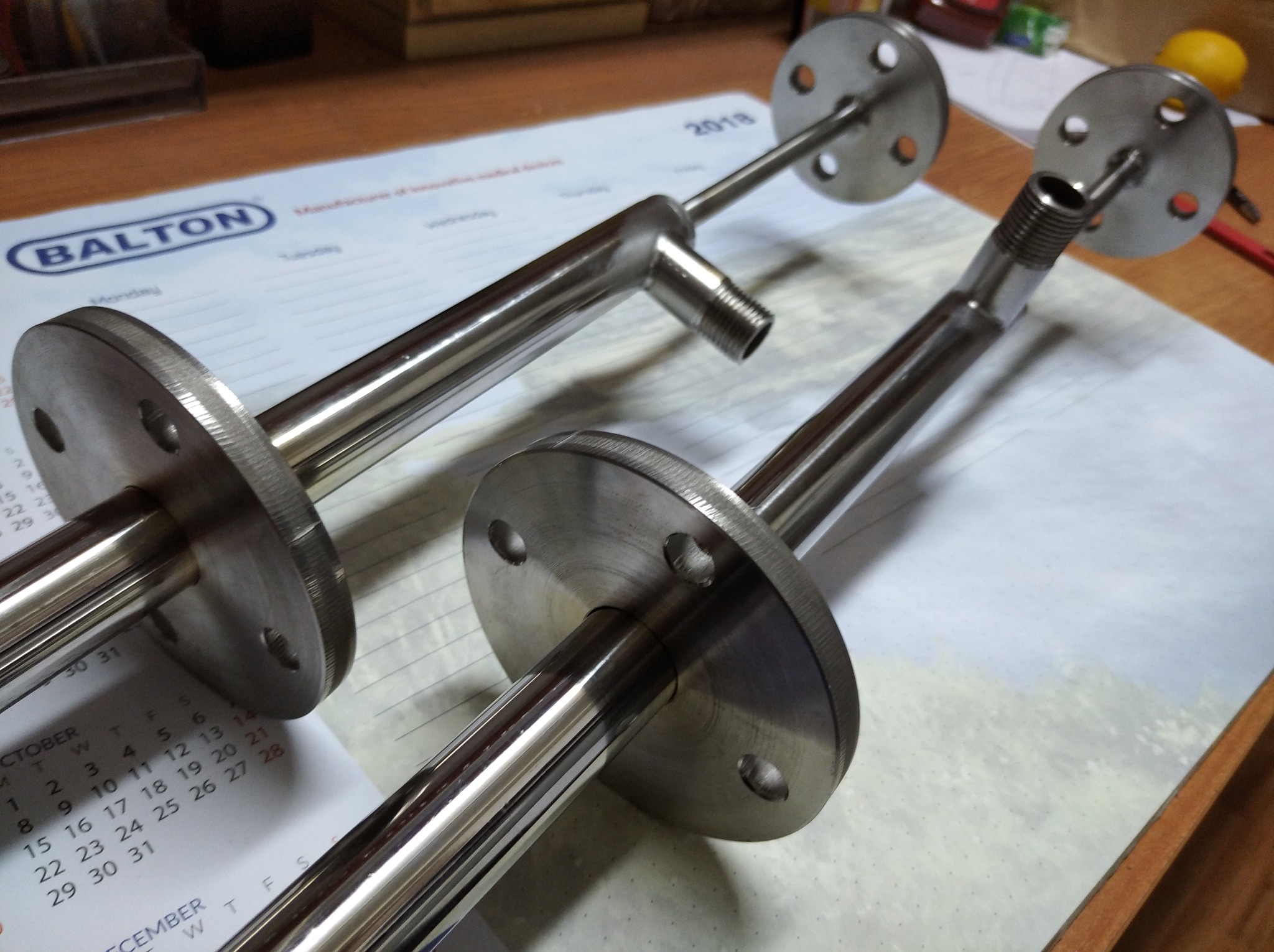 Dwa elementy metalowe z kołnierzami i gwintowanymi końcówkami na kalendarzu. Precyzyjna obróbka, widoczne ślady toczenia. Elementy wykonane z polerowanej stali.