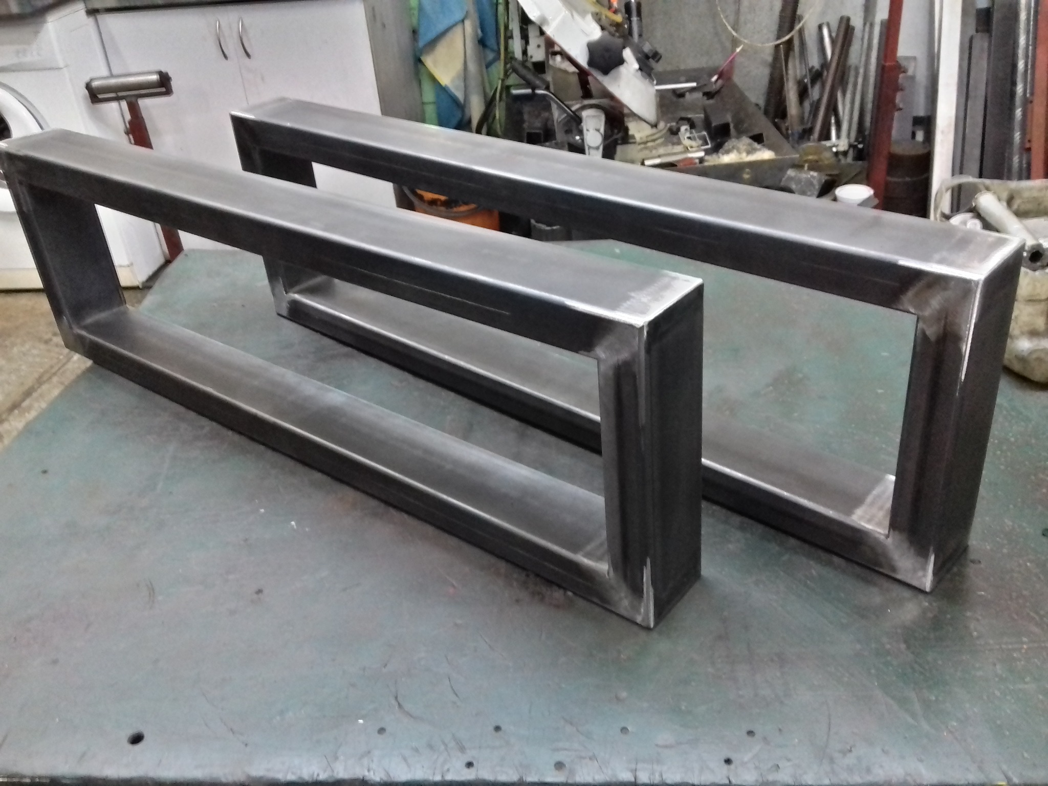 Dwa prostokątne, metalowe stelaże spawane z profili, leżące na warsztatowym stole. Widoczne ślady spawania i obróbki metalu. Tło warsztatowe z narzędziami.