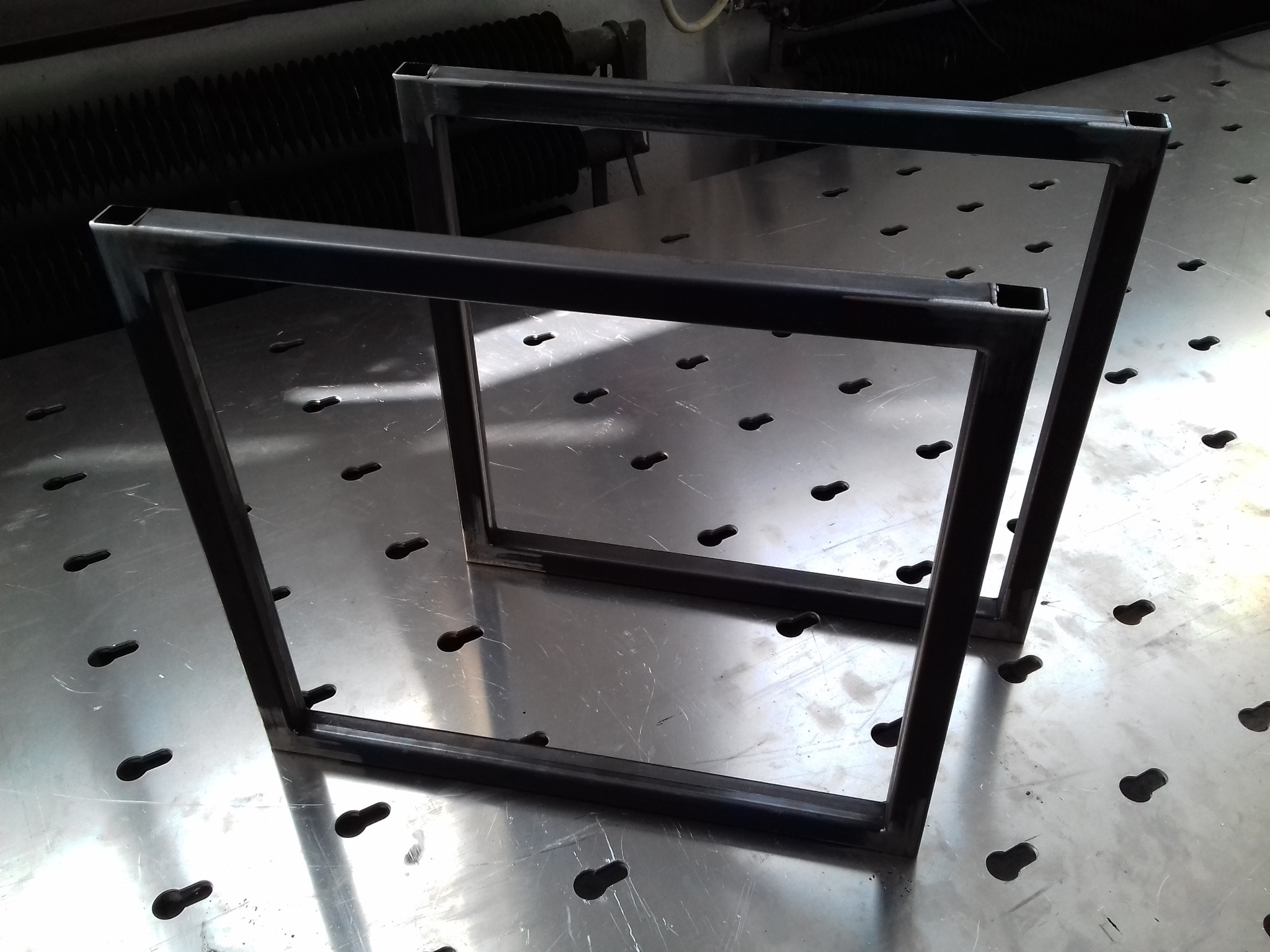 Dwie spawane ramy z profili kwadratowych na metalowym stole spawalniczym z otworami. Widoczne spawy na rogach. Surowy metal, gotowe elementy konstrukcyjne.