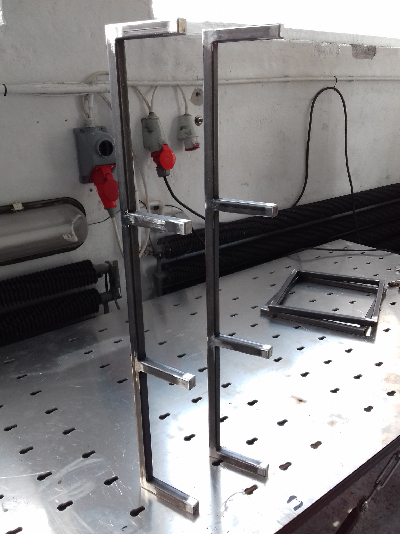 Dwa metalowe stelaże z przyspawanymi poprzeczkami na perforowanym stole warsztatowym. W tle gniazda elektryczne i fragmenty radiatora.