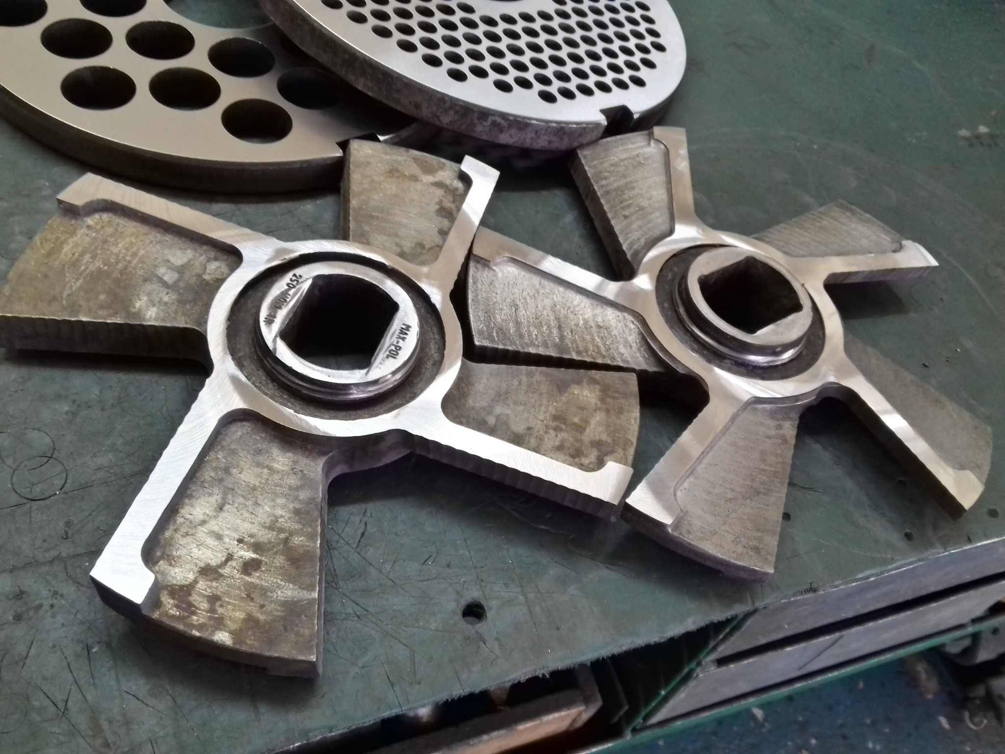 Dwa noże krzyżowe do maszynki do mielenia mięsa, obok tarcza z otworami, leżące na zielonym, metalowym blacie. Widoczne ślady obróbki.