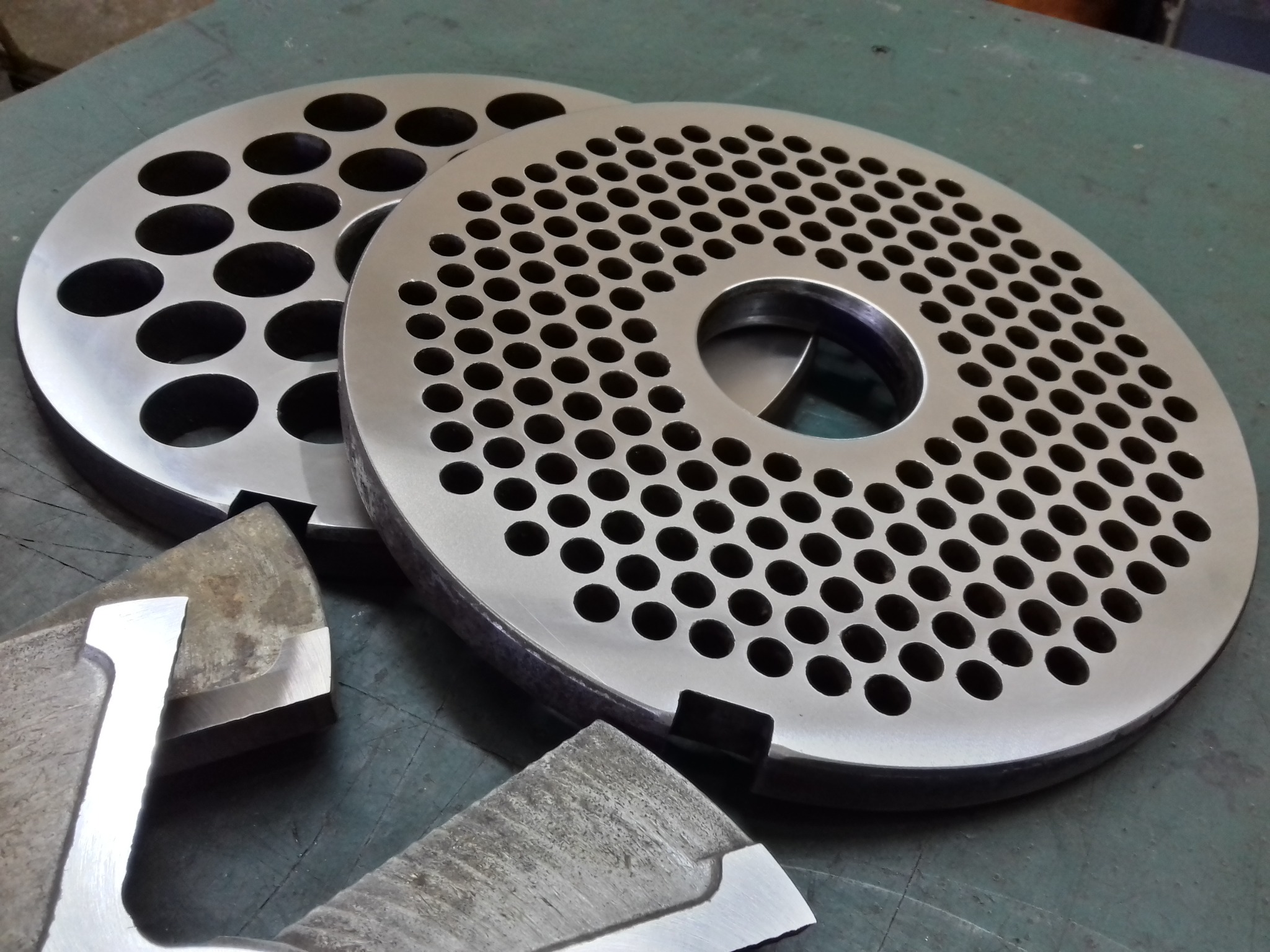 Dwie metalowe tarcze z otworami o różnej średnicy i noże do maszynki do mielenia mięsa na tle zielonego blatu warsztatowego.