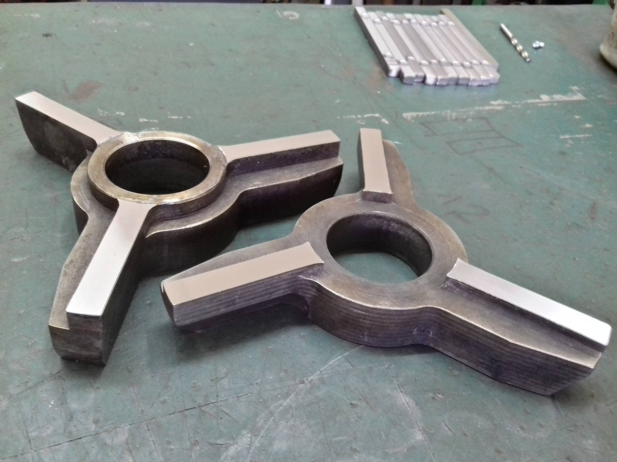 Dwa metalowe, symetryczne elementy z otworem centralnym i trzema ramionami, leżące na zielonym stole warsztatowym obok narzędzi. Obróbka metalu.