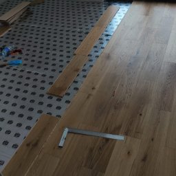 MONT-TRANS SPÓŁKA Z OGRANICZONĄ ODPOWIEDZIALNOŚCIĄ - Układanie jasnych paneli podłogowych w pokoju, widoczny kątownik i podkład. Częściowo ułożona podłoga w nowym mieszkaniu z oknem w tle.