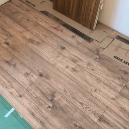 MONT-TRANS SPÓŁKA Z OGRANICZONĄ ODPOWIEDZIALNOŚCIĄ - Układanie paneli laminowanych w pomieszczeniu, widoczna podkładka pod panele i częściowo zamontowana drewniana ościeżnica drzwiowa.