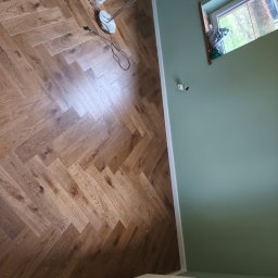 MONT-TRANS SPÓŁKA Z OGRANICZONĄ ODPOWIEDZIALNOŚCIĄ - Ukończona podłoga z parkietu ułożonego w jodełkę, widoczne narzędzia malarskie i grzejnik. Ściana pomalowana na zielono, widok z góry.