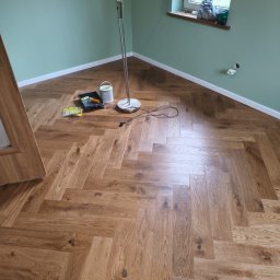 MONT-TRANS SPÓŁKA Z OGRANICZONĄ ODPOWIEDZIALNOŚCIĄ - Ukończona podłoga z parkietu ułożonego w jodełkę, widoczna lampa stojąca, puszka farby i akcesoria malarskie przy ścianie w odcieniach zieleni.