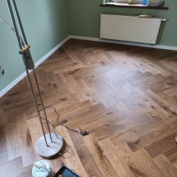 MONT-TRANS SPÓŁKA Z OGRANICZONĄ ODPOWIEDZIALNOŚCIĄ - Ukończona podłoga z paneli ułożonych w jodełkę, widok z góry, z farbą i narzędziami malarskimi w rogu. Ściany w kolorze zielonym, okno z grzejnikiem.