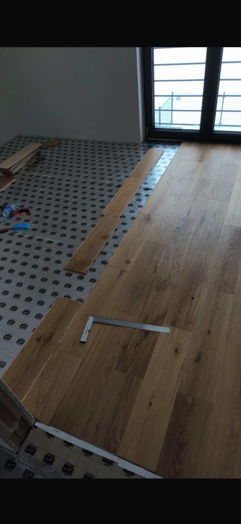 Układanie jasnych paneli podłogowych w pokoju, widoczny kątownik i podkład. Częściowo ułożona podłoga w nowym mieszkaniu z oknem w tle.