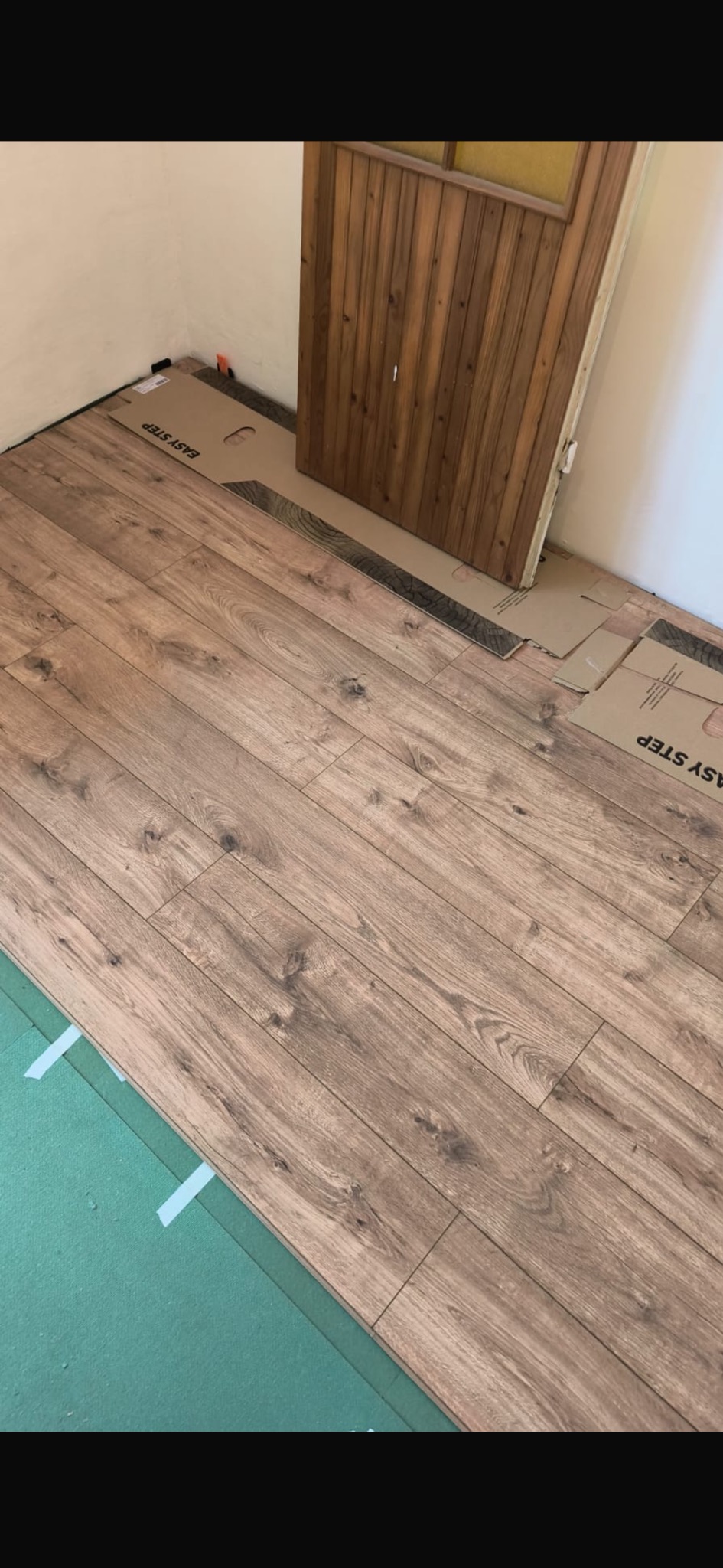 Układanie paneli laminowanych w pomieszczeniu, widoczna podkładka pod panele i częściowo zamontowana drewniana ościeżnica drzwiowa.