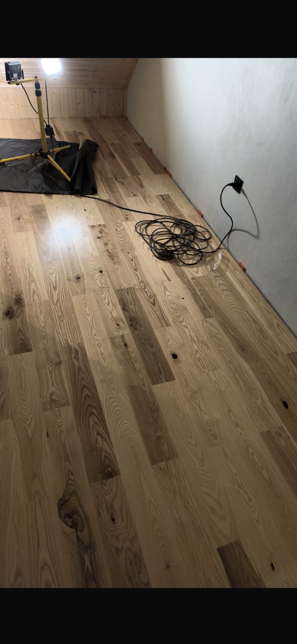 Ułożona podłoga z desek w jasnym odcieniu drewna, widoczne narzędzia pracy: lampa oświetleniowa i przewody elektryczne. Pomieszczenie w trakcie prac wykończeniowych.