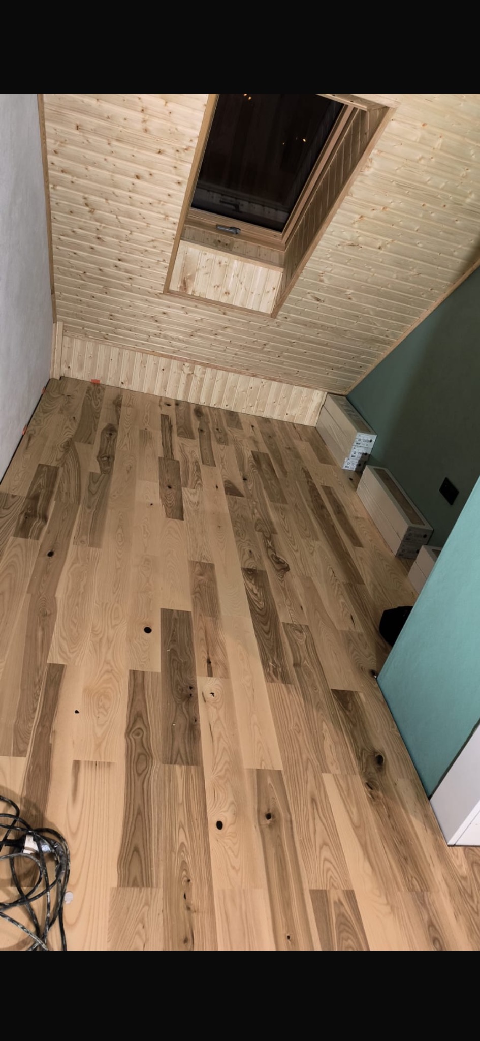 Jasna, drewniana podłoga z widocznymi otworami, ułożona w nowym pomieszczeniu z drewnianym sufitem i oknem dachowym. Widoczne kable w rogu.