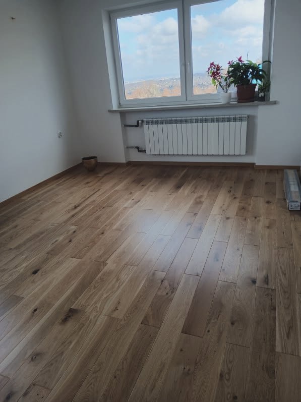 Jasna, drewniana podłoga ułożona w pokoju z grzejnikiem pod oknem. Widoczne detale usłojenia drewna i naturalne światło wpadające przez okno.
