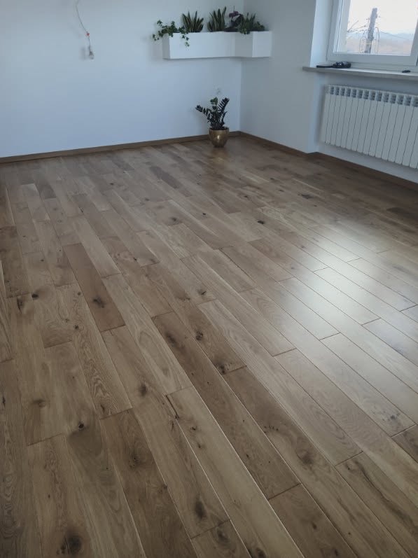 Jasne, drewniane panele podłogowe ułożone w pokoju z białymi ścianami, grzejnikiem i roślinami. Widok na całą podłogę, ukazujący wzór i kolorystykę.