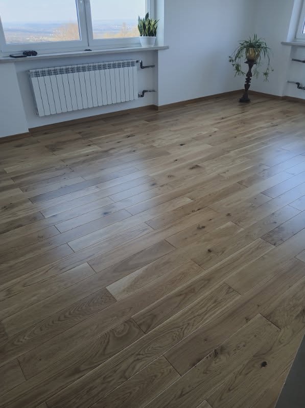 Jasna, drewniana podłoga ułożona w jodełkę, widok z góry, z grzejnikiem i roślinami w tle. Naturalne światło podkreśla strukturę drewna.