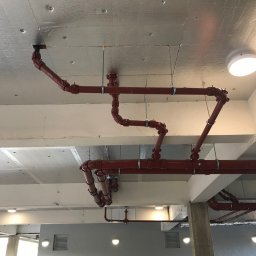 Hydro-Plumbing Ltd - Czerwone rury instalacji hydraulicznej pod sufitem, widoczne mocowania i łączenia, metaliczne wykończenie sufitu, oświetlenie sufitowe.