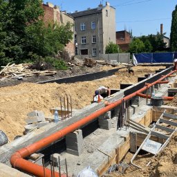 Hydro-Plumbing Ltd - Montaż pomarańczowych rur kanalizacyjnych w wykopie fundamentowym, widoczny pracownik przy łączeniu, częściowo zalane fundamenty, teren budowy z drewnem i piaskiem.